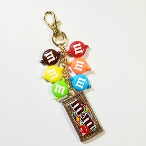 May include: A gold-toned metal keychain featuring a miniature rectangular chocolate bar charm with the M&M's logo and six round, colorful beads. Each bead is printed with a white lowercase m, including red, orange, yellow, green, blue, and brown.