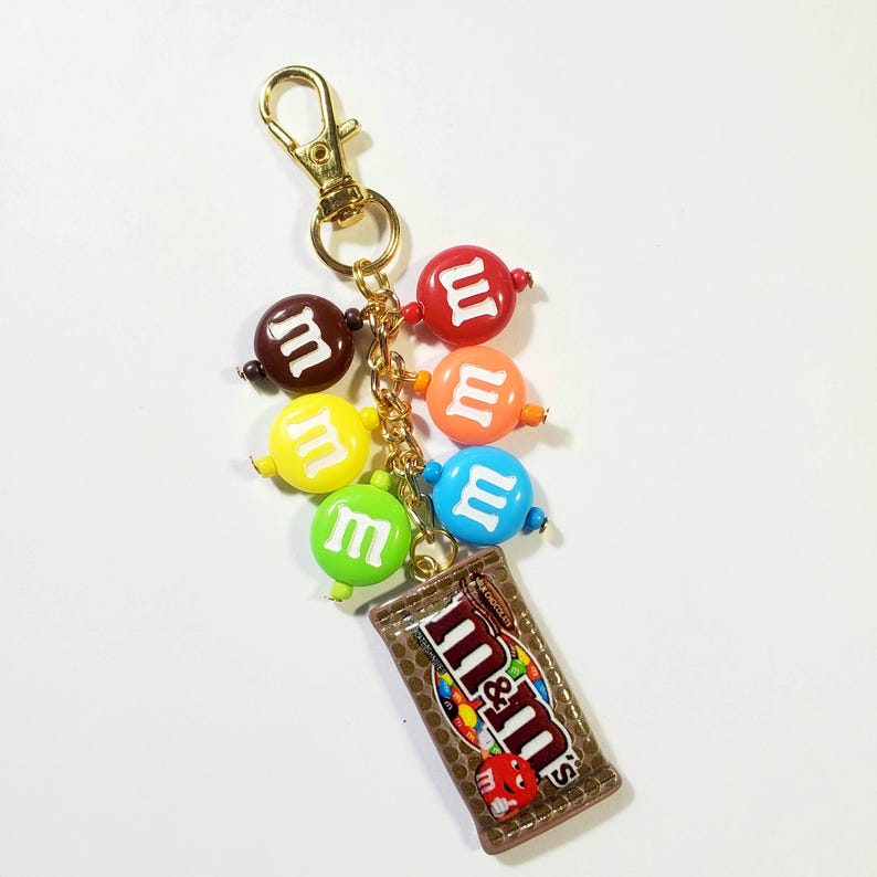 May include: A gold-toned metal keychain featuring a miniature rectangular chocolate bar charm with the M&M's logo and six round, colorful beads. Each bead is printed with a white lowercase m, including red, orange, yellow, green, blue, and brown.