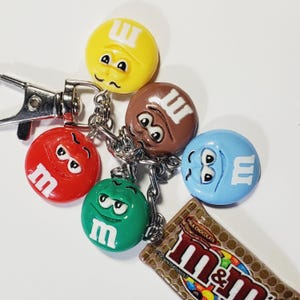 May include: A metal keychain featuring five charms: four round, colorful M&M character faces in yellow, brown, red, green, and blue, and one rectangular charm depicting a Milk Chocolate M&M candy bar wrapper with the text M&M's visible.