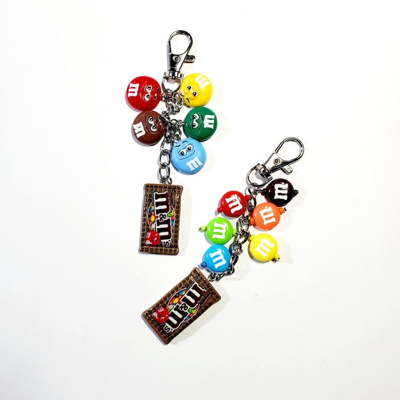 May include: Two silver-toned metal keychains featuring miniature colorful candy-shaped charms and a rectangular brown candy bag charm with the text m&m's printed on it. The charms are attached to a silver chain with a lobster claw clasp.