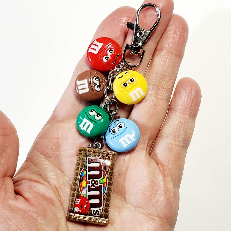 May include: A metal keychain featuring a silver lobster clasp attached to a chain with five charms. Four charms are round, colored red, brown, yellow, green, and blue, each with a white letter m. One rectangular charm depicts an M&M's candy bag.