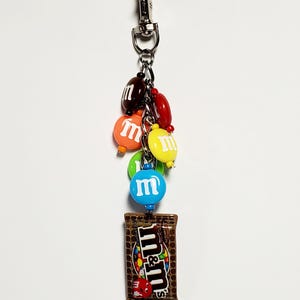 May include: A silver-toned metal lobster clasp keychain featuring a collection of colorful, round, candy-shaped charms in red, orange, yellow, green, blue, and brown, each with a white letter m, and a rectangular brown candy bag charm with m&m's text.