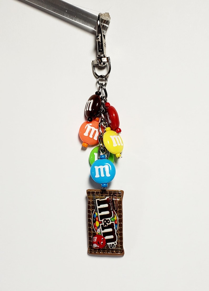 May include: A silver-toned metal lobster clasp keychain featuring a collection of colorful, round, candy-shaped charms in red, orange, yellow, green, blue, and brown, each with a white letter m, and a rectangular brown candy bag charm with m&m's text.