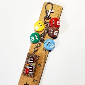 May include: A metal keychain featuring five colorful M&M character charms in red, yellow, green, brown, and blue, plus a rectangular M&M's logo charm. The keychain is displayed against a wooden ruler showing measurements up to 5 inches in length.