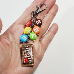 May include: A keychain featuring a silver-toned metal lobster clasp attached to a chain with several small, colorful, round candy-shaped charms in red, yellow, blue, green, orange, and brown, each with a white letter m, and a rectangular charm.