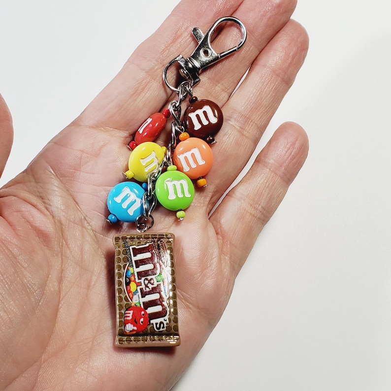 May include: A keychain featuring a silver-toned metal lobster clasp attached to a chain with several small, colorful, round candy-shaped charms in red, yellow, blue, green, orange, and brown, each with a white letter m, and a rectangular charm.