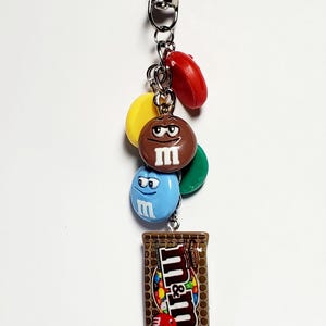 May include: A metal keychain featuring a silver lobster clasp and a collection of charms including red, yellow, green, brown, and blue M&M candy-shaped pieces with faces, and a rectangular charm depicting a brown M&M's milk chocolate candy bar.