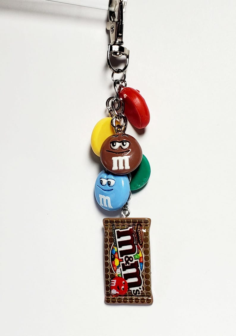 May include: A metal keychain featuring a silver lobster clasp and a collection of charms including red, yellow, green, brown, and blue M&M candy-shaped pieces with faces, and a rectangular charm depicting a brown M&M's milk chocolate candy bar.