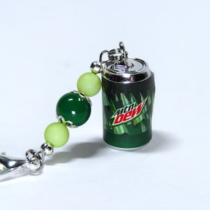 May include: A green and silver keychain featuring a miniature Mountain Dew can with a green and white striped design and the text "Mountain Dew". The keychain also includes a silver chain and green and light green beads.
