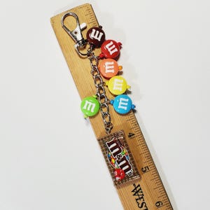 May include: A metal keychain featuring a silver lobster clasp and a chain adorned with five round, colorful beads resembling M&M candies in brown, red, orange, yellow, and blue, plus a rectangular charm with an M&M logo, measuring 6 inches long.