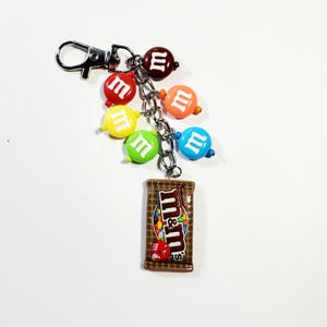 May include: A silver-toned metal keychain featuring a miniature brown M&M's chocolate candy bag charm and five round, colorful beads printed with the white letter m. The beads are red, yellow, green, blue, and orange, attached by a metal chain.