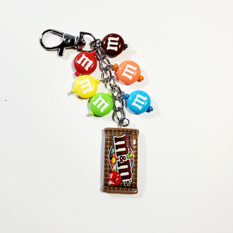 May include: A silver-toned metal keychain featuring a miniature brown M&M's chocolate candy bag charm and five round, colorful beads printed with the white letter m. The beads are red, yellow, green, blue, and orange, attached by a metal chain.