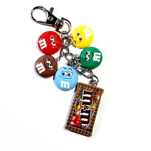 May include: A silver-toned metal keychain featuring a collection of five colorful M&M character charms in red, yellow, green, brown, and blue, along with a rectangular charm depicting a brown M&M's milk chocolate candy bar package with text.