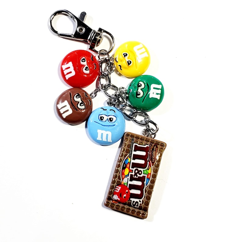 May include: A silver-toned metal keychain featuring a collection of five colorful M&M character charms in red, yellow, green, brown, and blue, along with a rectangular charm depicting a brown M&M's milk chocolate candy bar package with text.