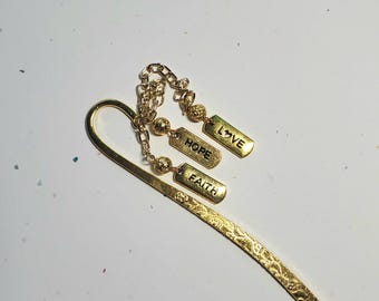 May include: A gold-colored metal bookmark with a textured design. The bookmark features three rectangular charms that read "LOVE", "HOPE", and "FAITH". The charms are connected to the bookmark with a delicate chain and beads.