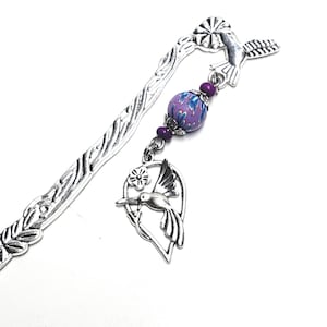 May include: A silver-toned metal bookmark with a hummingbird and floral design. The bookmark features a beaded accent with a purple and blue floral pattern. The hummingbird charm is in flight, adding a touch of nature.