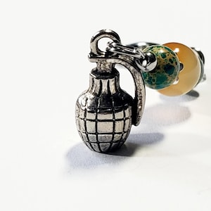 May include: A silver-colored grenade charm hangs from a ring with colorful beads. The grenade has a detailed, textured surface. The beads are green, yellow, and orange. The background is white.