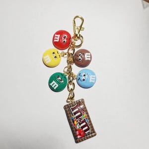 May include: A gold-toned metal keychain featuring five circular charms shaped like colorful M&M characters in red, yellow, brown, green, and blue, each with a white letter m. A rectangular charm depicting a brown M&M candy bag is attached.