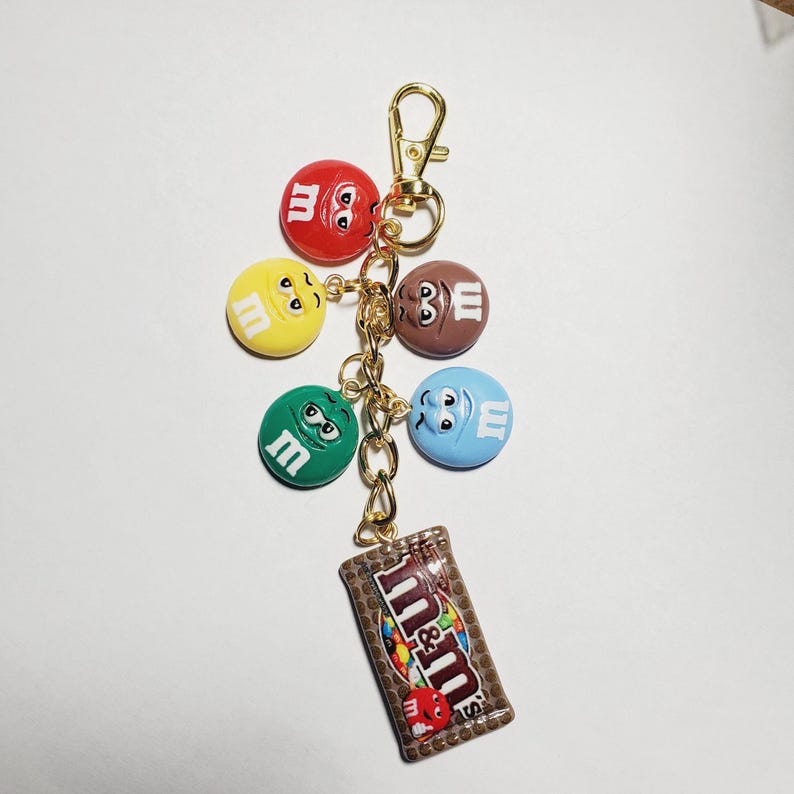May include: A gold-toned metal keychain featuring five circular charms shaped like colorful M&M characters in red, yellow, brown, green, and blue, each with a white letter m. A rectangular charm depicting a brown M&M candy bag is attached.