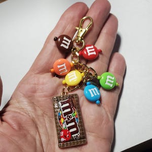 May include: A gold-toned metal keychain featuring a rectangular charm shaped like an M&M's chocolate bar package, accompanied by six round, colorful beads in brown, red, orange, yellow, green, and blue, each embossed with a white letter m.