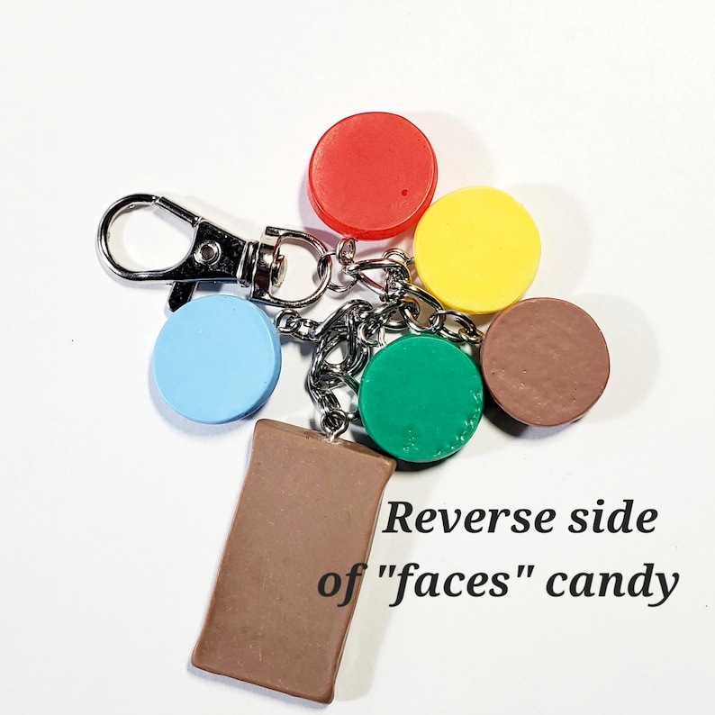 May include: A metal keychain featuring a silver lobster clasp and multiple charms, including five small solid-colored circles in red, yellow, brown, green, and light blue, plus a rectangular brown charm labeled Reverse side of "faces" candy.