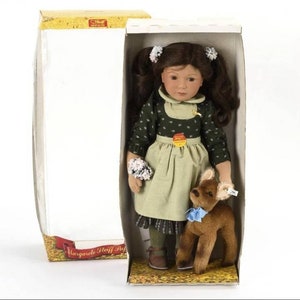 May include: A brown-haired doll wearing a green dress with a white apron and holding a bouquet of white flowers. The doll is standing next to a brown stuffed deer with a blue bow. The doll and deer are in a box with the text "Margarete Steiff Puppen" on the bottom.
