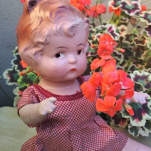 May include: A vintage porcelain doll with a red and white checkered dress. The doll has a painted face with brown hair and blue eyes. The doll is sitting in a garden setting with red flowers.