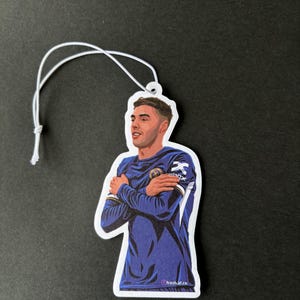 May include: A white car air freshener with a blue and white illustration of a soccer player with his arms crossed. The player is wearing a blue jersey with white accents and a white logo on the chest. The text "fresh.af.co" is printed on the bottom right corner of the air freshener.