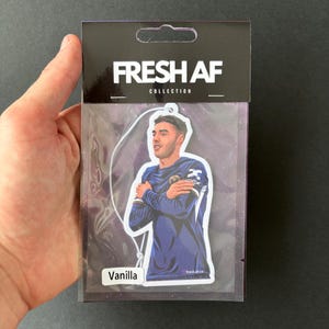 May include: A car air freshener with a photo of a soccer player on it. The air freshener is packaged in a clear plastic bag with a white header that says "FRESH AF COLLECTION" and a white label that says "Vanilla".
