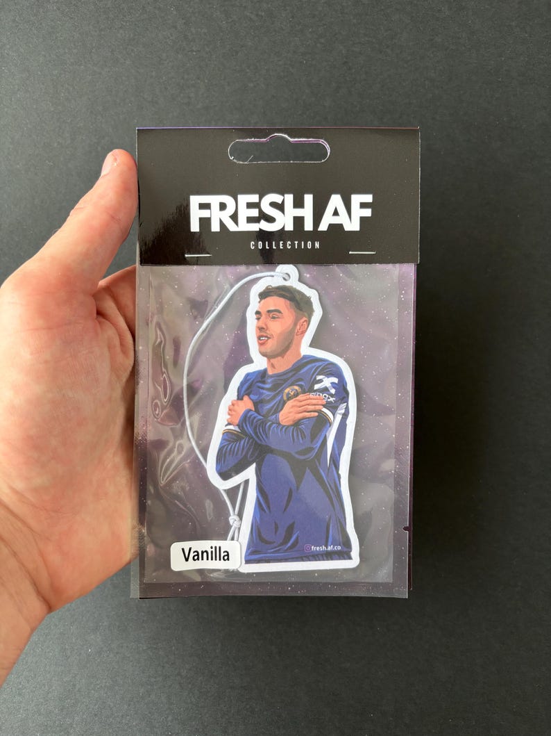 May include: A car air freshener with a photo of a soccer player on it. The air freshener is packaged in a clear plastic bag with a white header that says "FRESH AF COLLECTION" and a white label that says "Vanilla".