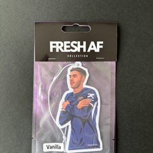 May include: A white car air freshener with a black border. The air freshener features a cartoon illustration of a soccer player in a blue jersey with the text "FRESH AF COLLECTION" at the top and "Vanilla" at the bottom.