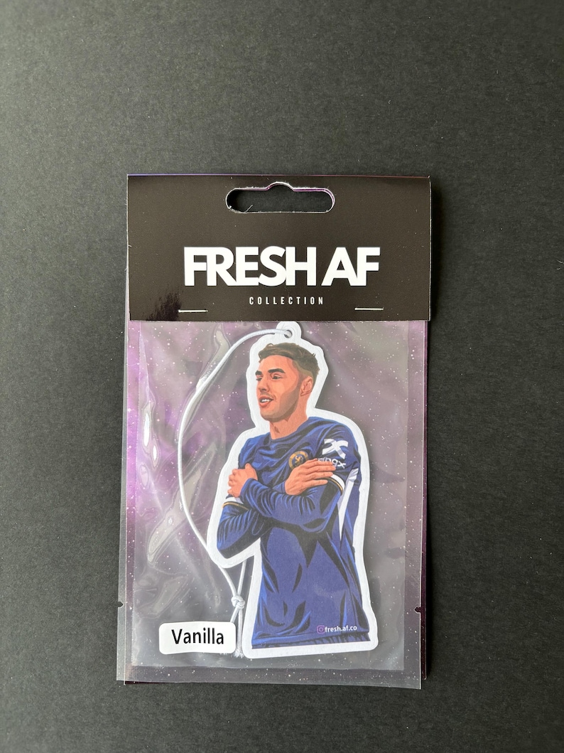 May include: A white car air freshener with a black border. The air freshener features a cartoon illustration of a soccer player in a blue jersey with the text "FRESH AF COLLECTION" at the top and "Vanilla" at the bottom.