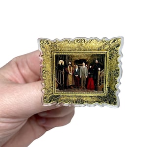 Puede incluir: Un pin de acrílico transparente con una imagen enmarcada de un grupo de personas. El marco dorado tiene detalles ornamentados. La imagen muestra a un grupo de personas en una escalera. El pin está sostenido por una mano.