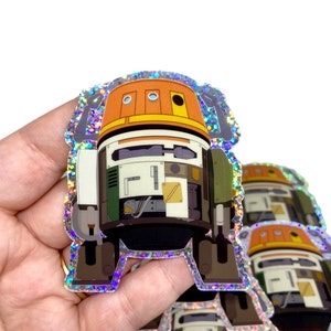 Op de afbeelding: Een holografische sticker van een robot met een oranje, wit en groen ontwerp. De robot heeft drie ronde ogen en een rechthoekig lichaam.