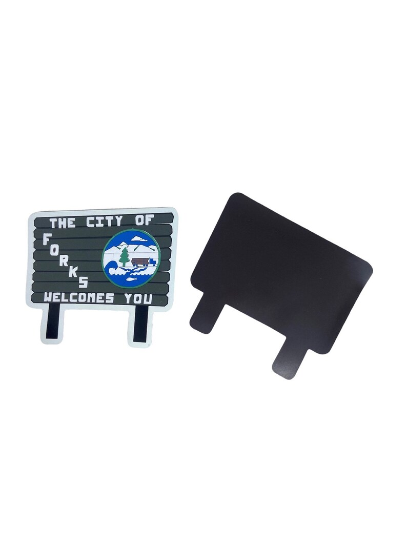 May include: A magnet featuring a sign that reads "THE CITY OF FORKS WELCOMES YOU." The sign has a gray background with white lettering and a blue and green graphic. A separate black magnet is also shown.