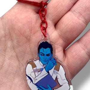 Pode incluir: Um chaveiro de acrílico transparente com um personagem de pele azul em uniforme branco, lendo um livro azul. O chaveiro tem uma corrente e um anel vermelhos. O personagem tem olhos vermelhos e está segurando o queixo.