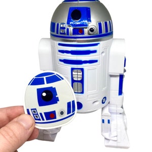 May include: A white and blue R2-D2 toy robot with a sticker of R2-D2 held in front. The robot has a silver head and blue accents. The sticker is white with blue details. The toy is a Star Wars collectible.