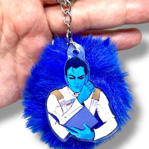 May include: A blue keychain featuring a character illustration with red eyes, holding a blue book, attached to a fluffy blue pom-pom. The keychain has a silver metal ring.