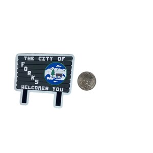 May include: A rectangular sign with the text "THE CITY OF FORKS WELCOMES YOU" in white letters. The sign features a circular emblem with a mountain, trees, and a cabin. A quarter is next to the sign.