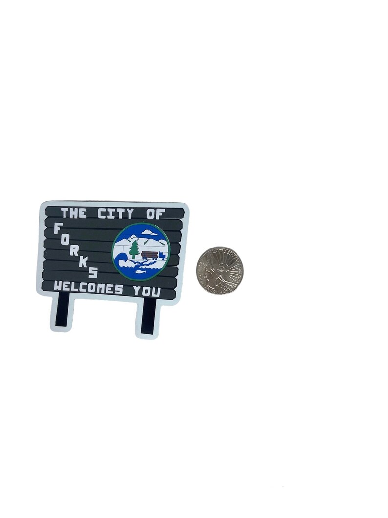 May include: A rectangular sign with the text "THE CITY OF FORKS WELCOMES YOU" in white letters. The sign features a circular emblem with a mountain, trees, and a cabin. A quarter is next to the sign.