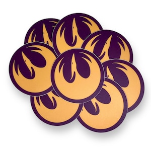 May include: A stack of eight round stickers featuring the Rebel Alliance symbol from Star Wars. The symbol is a white bird in flight with wings spread, on a gold background. The stickers have a purple border.