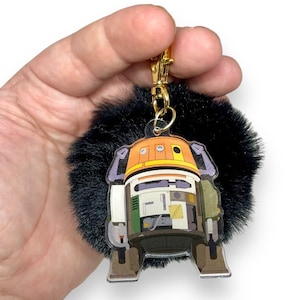 Op de afbeelding: Een zwarte, pluche sleutelhanger met een transparante acrylbedel met een cartoonrobot met oranje, witte en groene details.