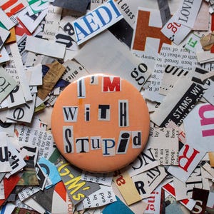 Può includere: Un bottone color pesca con le parole "I'm With Stupid" scritte in lettere nere ritagliate da pagine di riviste.