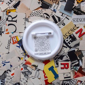 May include: A white button pin with a safety pin backing, featuring a QR code and the text "RAINY DAYS ARE OKAY." The pin is set against a background of torn newspaper clippings with various letters and colors.