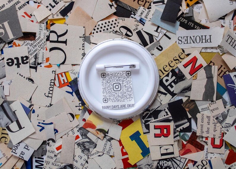 May include: A white button pin with a safety pin backing, featuring a QR code and the text "RAINY DAYS ARE OKAY." The pin is set against a background of torn newspaper clippings with various letters and colors.
