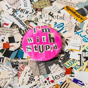 May include: A pink button with the words "I'm with Stupid" in black letters cut out from magazine pages. The button is surrounded by a variety of colorful magazine clippings.
