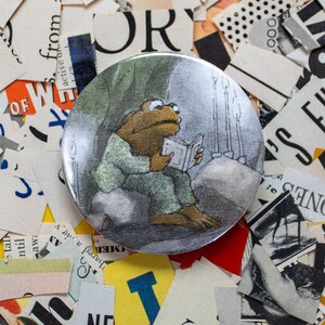 May include: A round button featuring an illustration of a frog reading a book. The frog is wearing a green outfit and is seated on a rock. The background is a dark, cave-like setting with candles. The button is surrounded by pieces of paper.