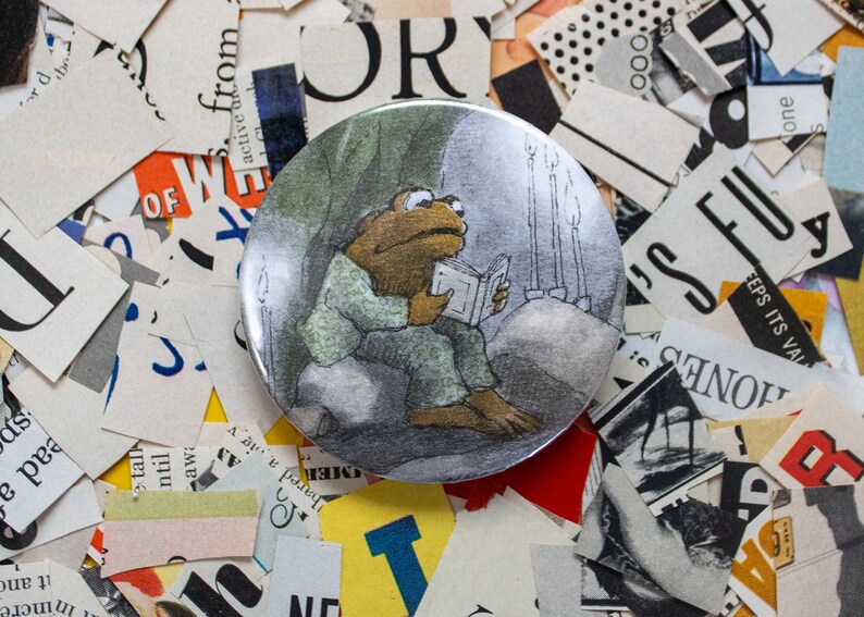 May include: A round button featuring an illustration of a frog reading a book. The frog is wearing a green outfit and is seated on a rock. The background is a dark, cave-like setting with candles. The button is surrounded by pieces of paper.
