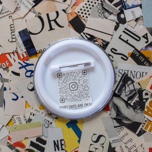 May include: A white button pin with a safety pin back, featuring a QR code and the text "RAINY.DAYS.ARE.OKAY". The pin is set against a background of torn paper with various printed text and colors.