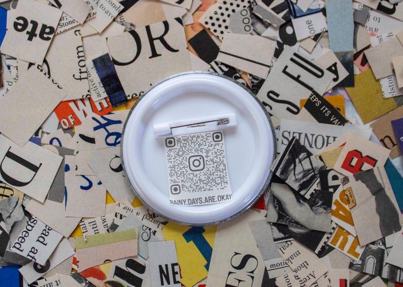 May include: A white button pin with a safety pin back, featuring a QR code and the text "RAINY.DAYS.ARE.OKAY". The pin is set against a background of torn paper with various printed text and colors.